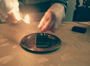 Psarah's hands striking match. A gold ashtray and matchbox are on a gold brocade tablecloth. 